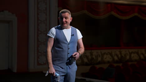 presenter on stage. man holding medical mask in his hand and talking on camera in empty theater. portrait of middle-aged man speaker. pandemic concept.