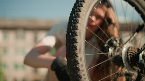 close-up of rear bicycle tire with rugged tread pattern inspected for air pressure by person wearing black gloves, fingers pressing firmly on tire sidewall, softly blurred building in background