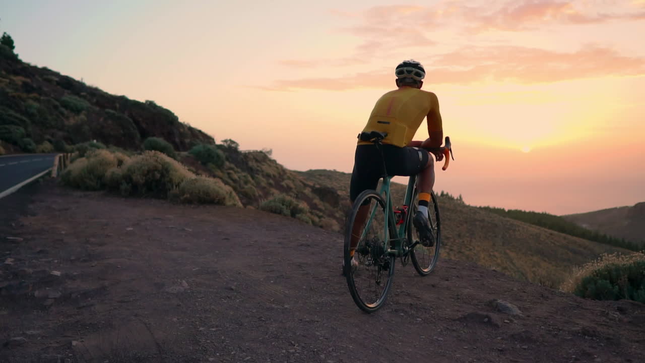 Post-training, athlete in yellow t-shirt, helmet, and gear sits on bike on mountain peak, enjoying view of mountains and sunset