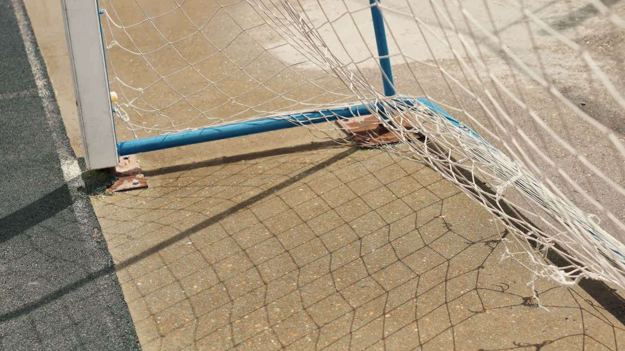 Primo piano di un palo bagnato e un pallone da calcio appoggiato sulla rete dopo una partita piovosa, con pavimento pozzangheroso, rete a maglie luccicante e palo dipinto di blu. L'ombra del campo vuoto e la luce nuvolosa creano un'atmosfera riflessiva e malinconica