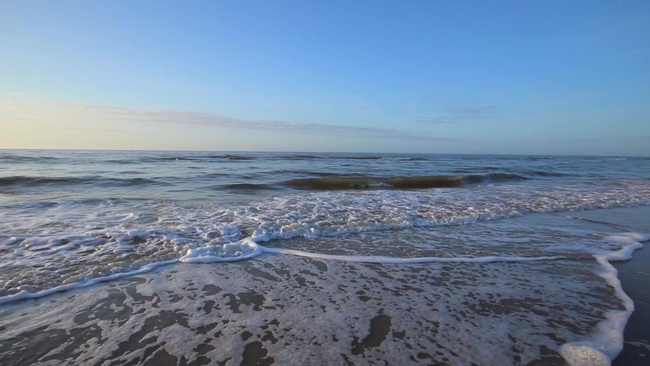Onde che si infrangono e risalgono la riva della spiaggia al mattino. Onde dell'oceano che si frangono sulla battigia. Bolle e schiuma sulle onde marine. Inquadratura grandangolare che si allontana. Movimento lento. Alba