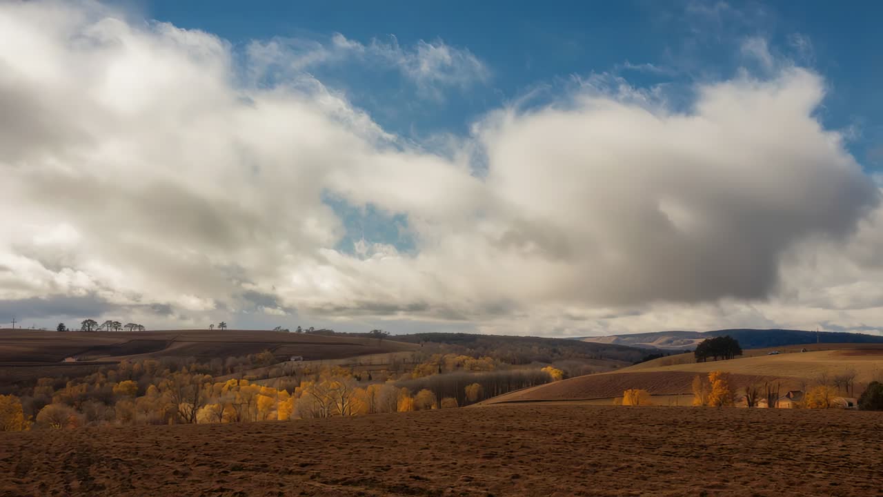 Drifting cumulus clouds moving across hillside, casting shadows over tilled field with fall foliage