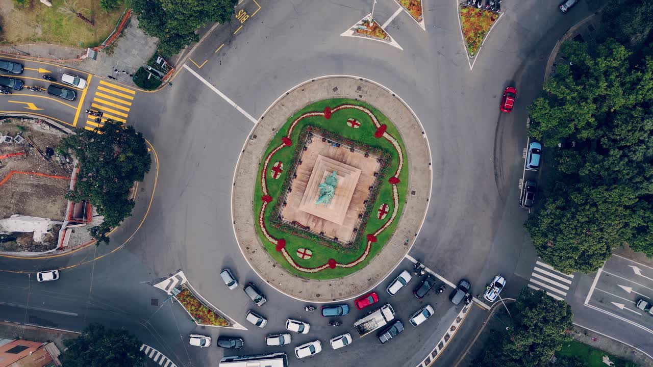 Cars navigate a busy roundabout in Genoa, Italy, surrounded by greenery and city streets