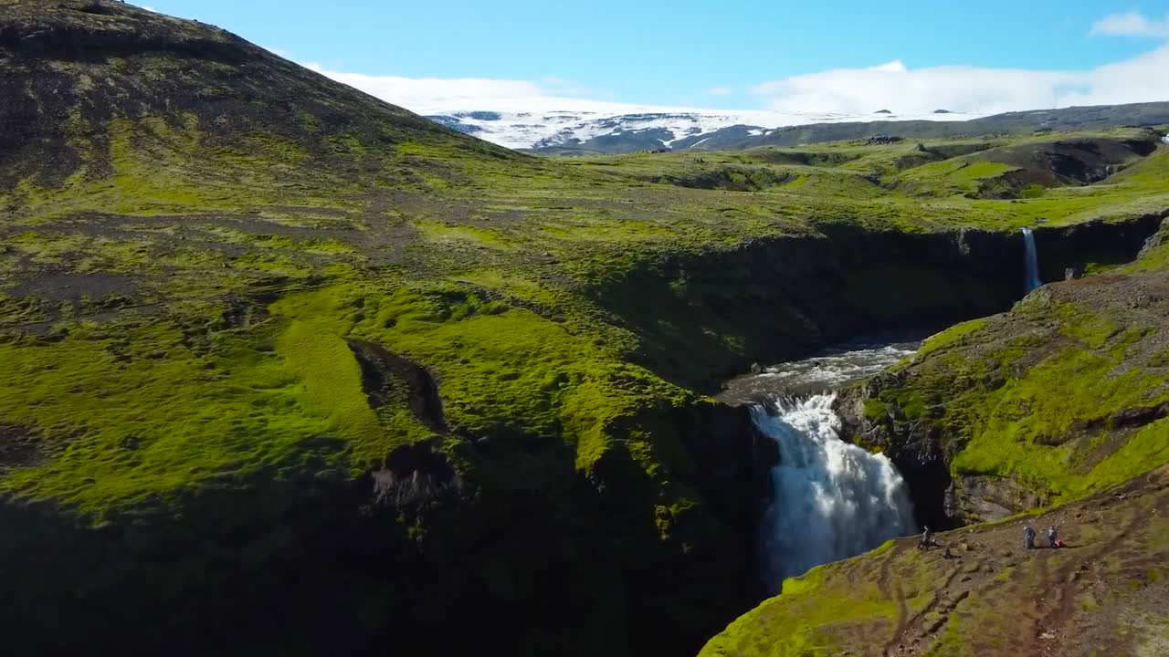 Aerial drone footage flying over and closer to a waterfall in Iceland that has fast flowing river and white water flowing through mossy green and dark brown rocky volcanic terrain during a sunny day.