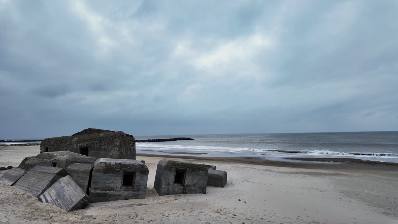 The remains of a World War II bunker stand silently on the beach by the sea, as the calm waters stretch endlessly beneath a cloudy sky.