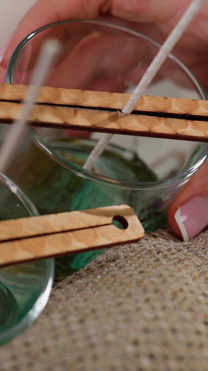 Primo piano di mani che usano un attrezzo per stoppini in legno per intingere uno spago di cotone in cera verde per la creazione di candele. Le mani tengono un contenitore di vetro con cera colorata fusa e un attrezzo, concentrandosi su una lavorazione precisa.