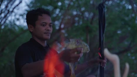a happy spear fisherman taking two fish that he has caught off his speargun with a fire burning in front of him ready to cook his fish