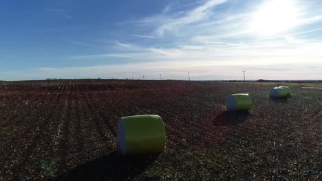 L-to-R-panning-Drone-Aerial-shot-with-a-dramatic-rise-to-the-sun-over-a-Midwestern-cotton-farm-with-fresh-bales-of-harvested-cotton-wrapped-in-bright-yellow-material-against-a-blue-open-sky