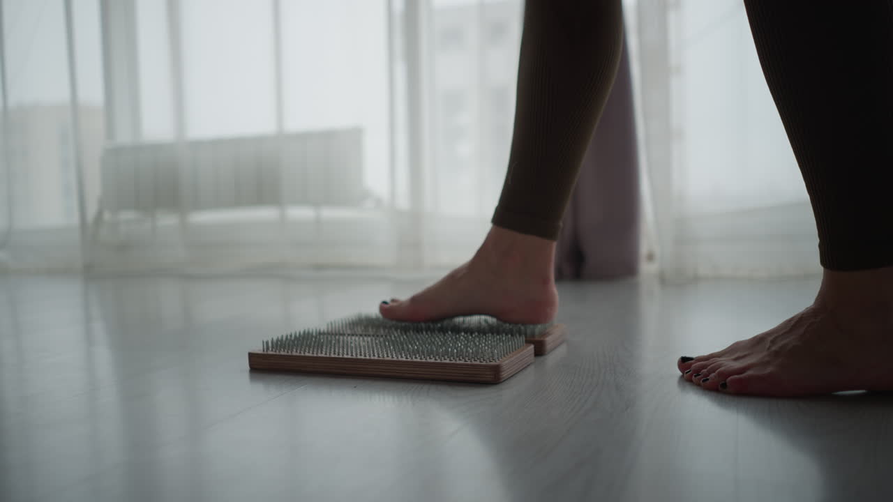 Graceful woman lowers barefoot onto wooden nail board on pink yoga mat in modern studio with red aerial silks and floor to ceiling windows during mindful balance practice for wellness training