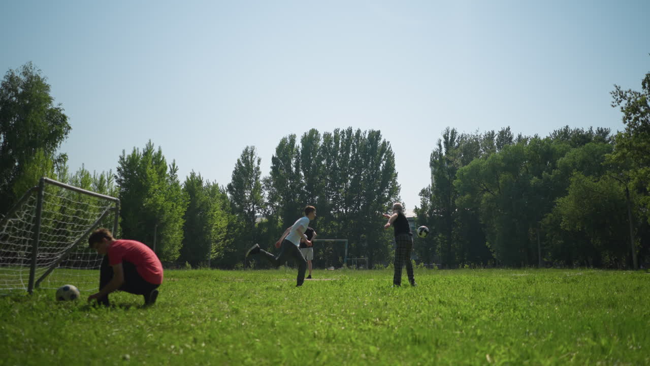 A grandfather and grandson play football, the grandfather throws, the grandson heads the ball, nearby, someone ties his shoelace near a goalpost