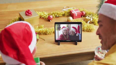 Kaukasischer-Vater-Und-Sohn-Mit-Weihnachtsmützen-Nutzen-Tablet-Für-Weihnachtsvideoanruf-Mit-Paar-Auf-Dem-Bildschirm