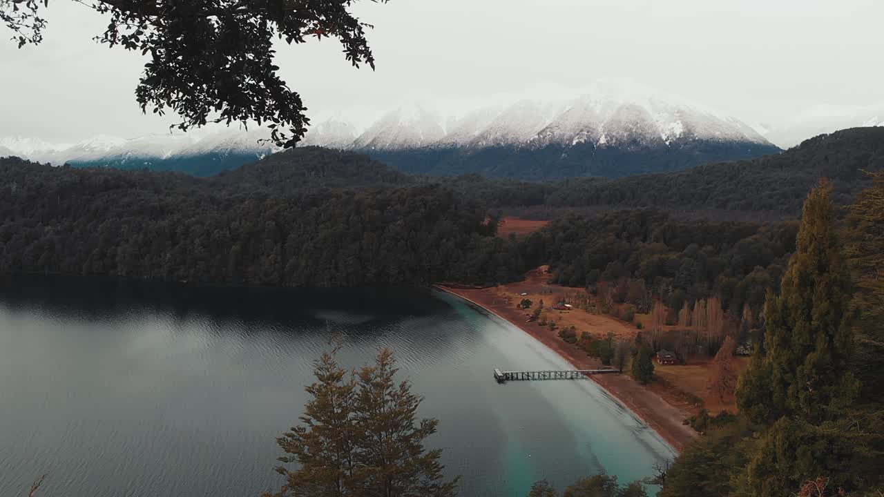 vista desde un mirador a la orilla de un lago con un muelle, un bosque y montañas nevadas al fondo