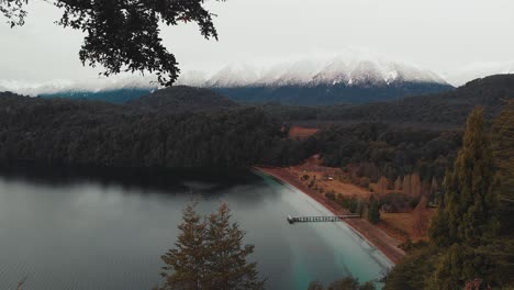 view from a viewpoint to the shore of a lake with a pier, forest and snowy mountains in the background