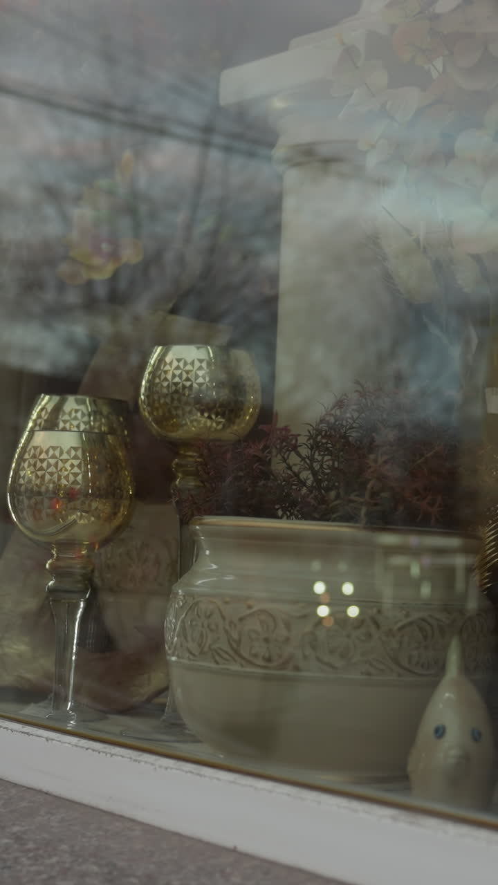 vista de cerca de una vitrina de una tienda con elegantes artículos decorativos, incluidas copas de vidrio ornamentadas, ollas de cerámica y plantas, con reflexión en el vidrio