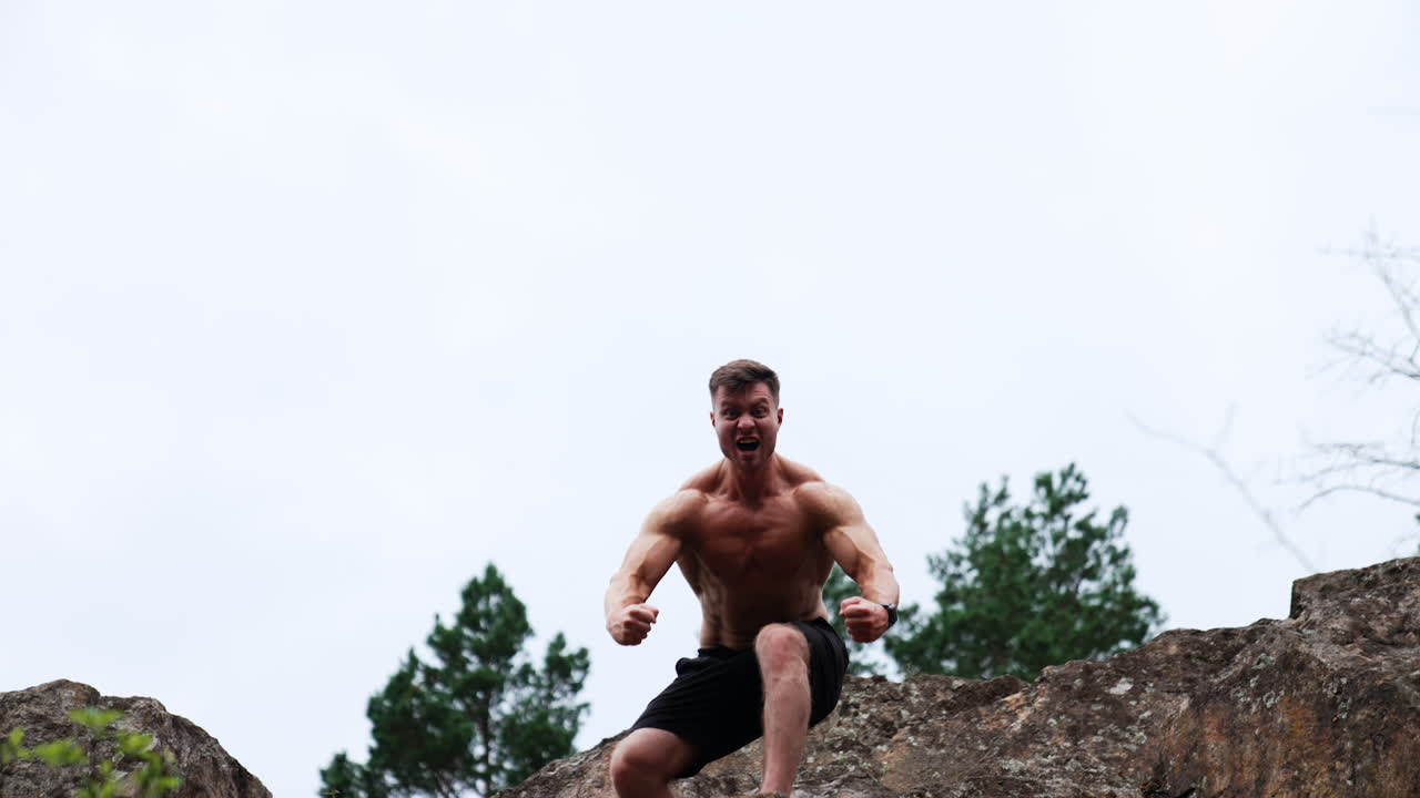 Caucasian strongman demonstrating his muscles standing on the rock. Athlete strains his arms and screams. Low angle view.