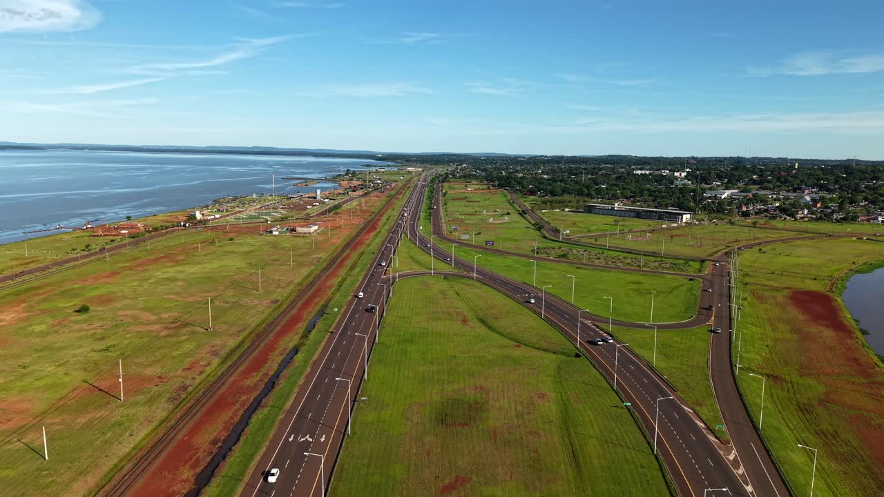 Aerial follow of Acceso Sur highway in Posadas, Misiones, Argentina, with multiple lanes, sweeping curves, and green open embankments running beside the wide Paraná River and connecting to the city