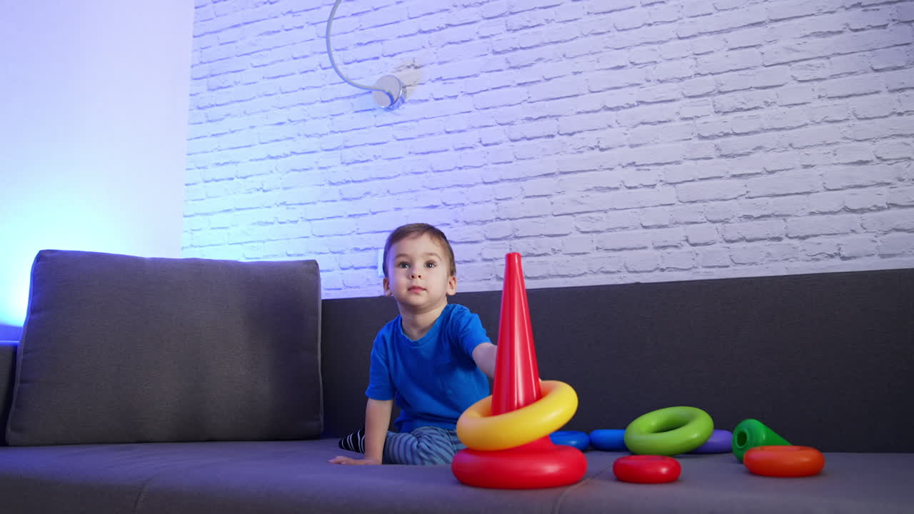 Cute dark-haired toddler assembling pyramid on the sofa. Baby boy plays with toy at home.