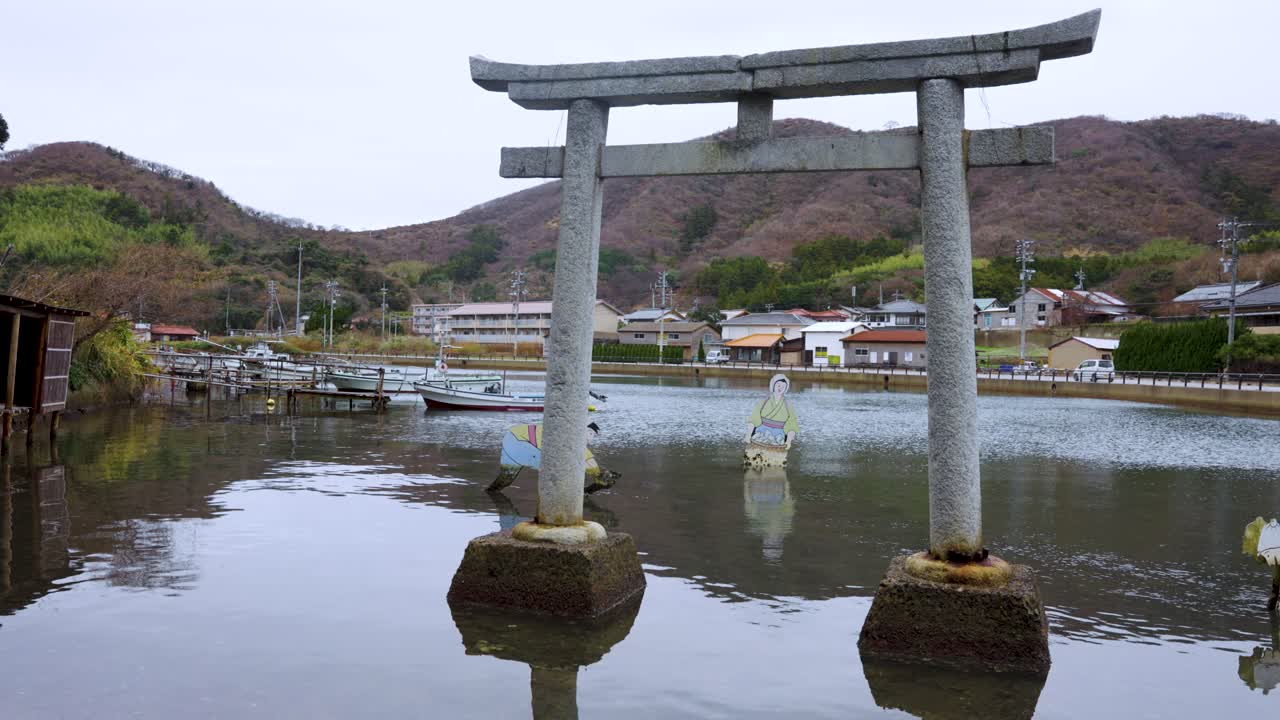 Tradiční japonská brána Torii stojící ve vodě s vesnicí a kopci