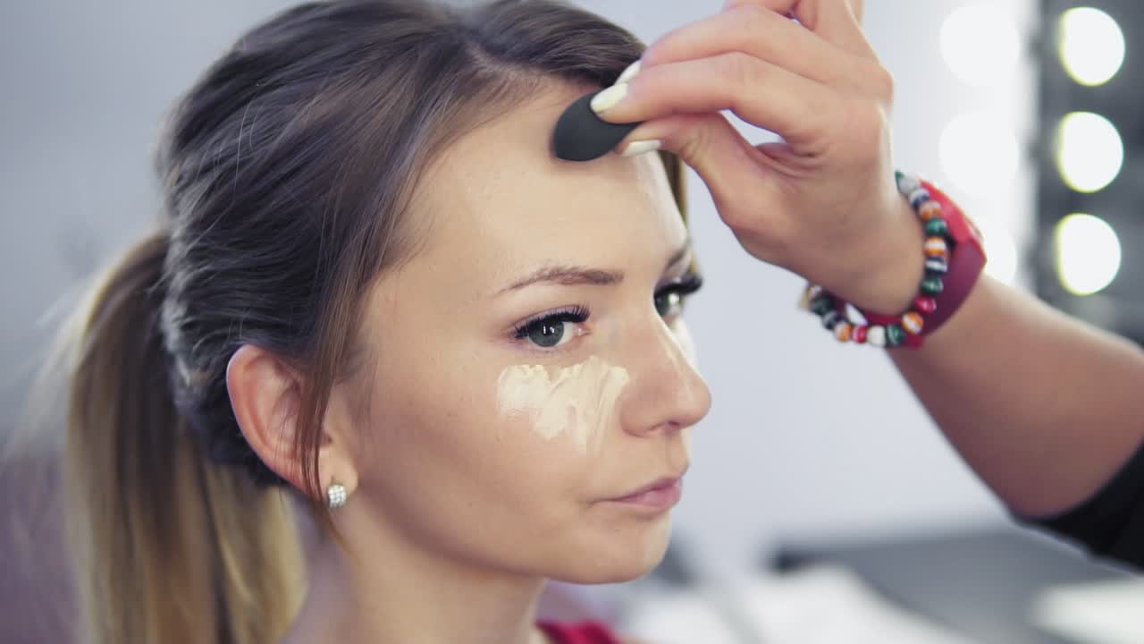 primer plano de un artista de maquillaje profesional aplicando corrector a la piel de una mujer joven con pincel para cubrir las ojeras y maquillar la cara