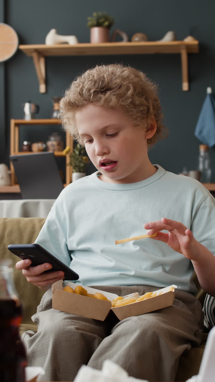 Young Boy Eating Fast Food and Using Smartphone on Couch