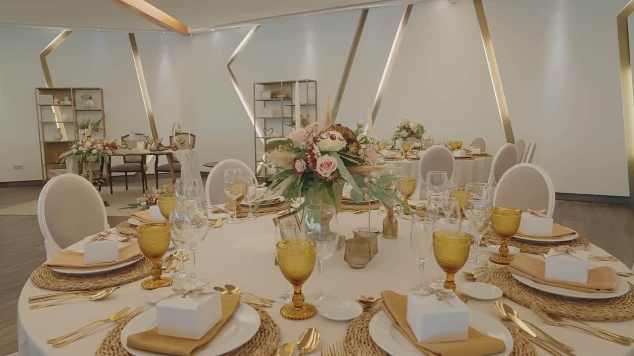 Runde borde dækket med guldbestik, ravfarvede glas, blomsteropsatser og hvide stole i en lys, moderne sal.