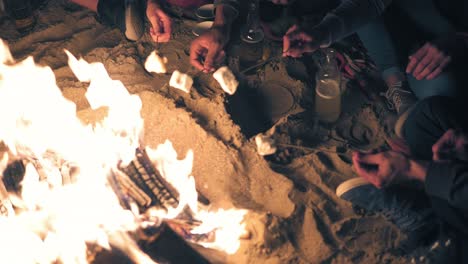 view from the top: hands holding sticks with marshmallows and frying them at night. group of people sitting by the fire late at night