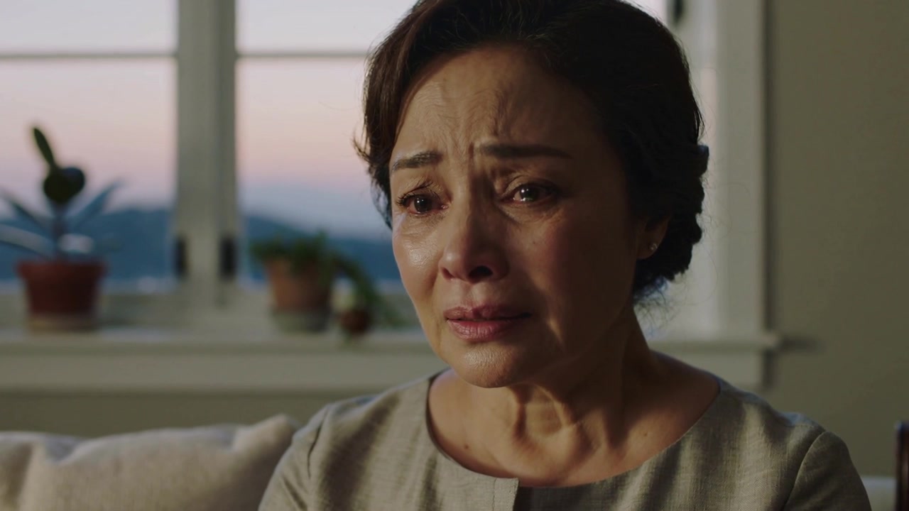 Crying senior Asian woman in grey blouse after looking off-screen on couch at home with plants