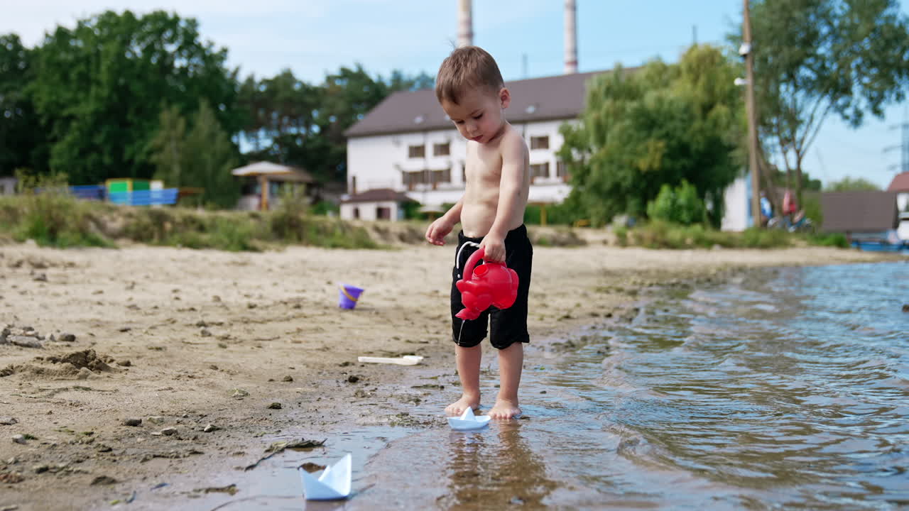 Adorable baby boy with a red watering can pouring water. Sweet child playing on the shore in summer.