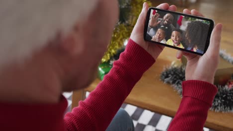 hombre caucásico con sombrero de santa usando un teléfono inteligente para una videollamada de navidad con una familia sonriente en la pantalla