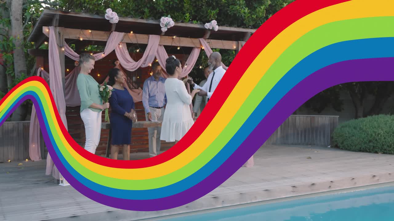 Woman in white dress reading vows at ceremony, man lifting ring, rainbow sweeping over couple
