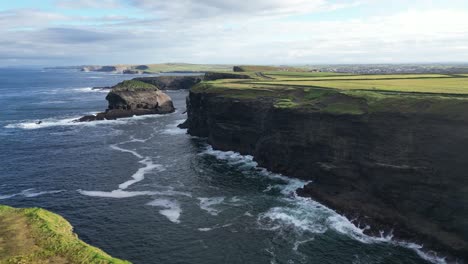 vista aérea de acantilados escarpados junto al mar con olas que se estrellan, campo verde en el fondo