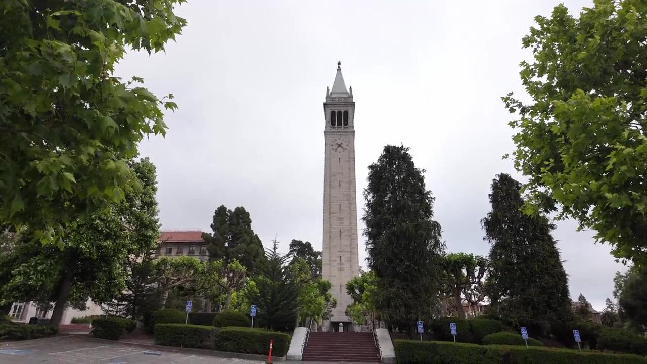 Una ripresa grandangolare stabile cattura la Sather Tower incorniciata da alberi di un verde vibrante in una giornata nuvolosa all'UC Berkeley, mettendo in mostra l'iconica architettura universitaria e dintorni tranquilli e vuoti.