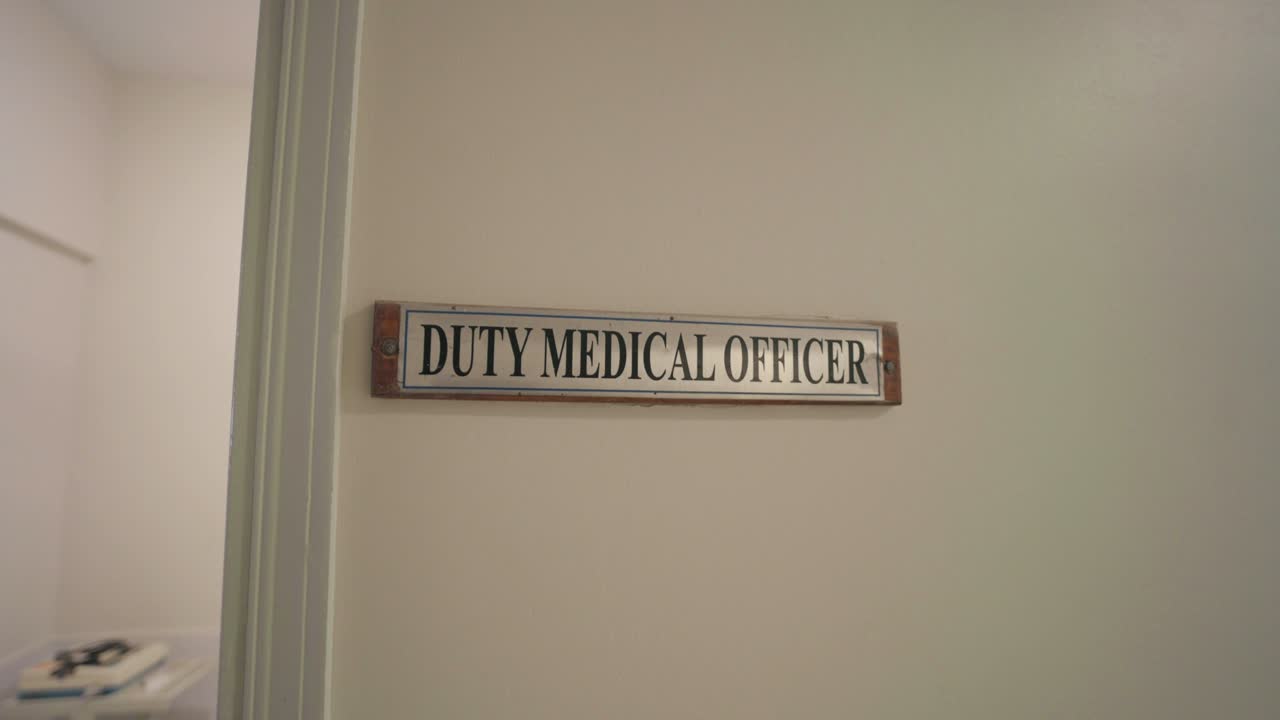 Sign reading "DUTY MEDICAL OFFICER" mounted beside a doorway on a cream-colored wall inside a hospital corridor, captured in a zoom-in movement with ambient daylight