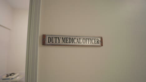 sign reading "duty medical officer" mounted beside a doorway on a cream-colored wall inside a hospital corridor, captured in a zoom-in movement with ambient daylight
