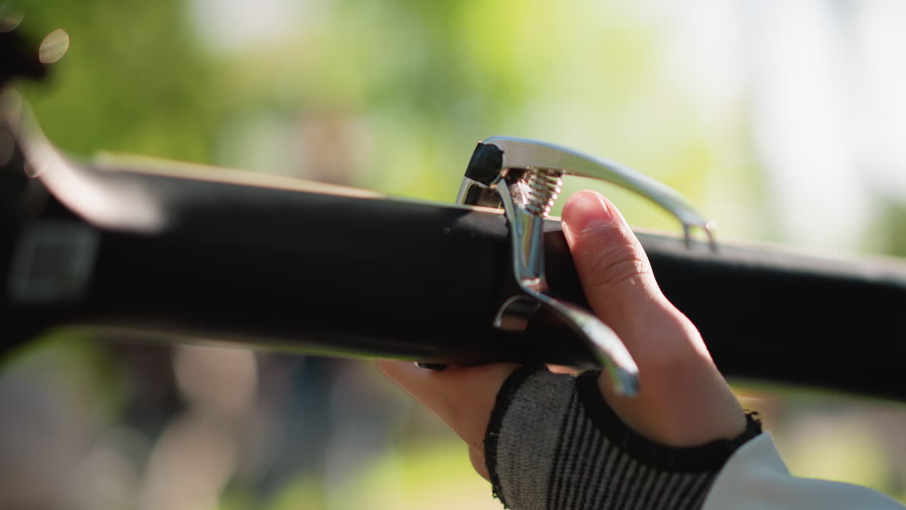 primo piano di una mano che posiziona un capotasto su una chitarra, luce solare in un parco all'aperto, polpastrello che regola il morsetto sul manico acustico, texture di un guanto senza dita, clic del morsetto che prepara per l'esibizione di strada, atmosfera da sessione di pratica
