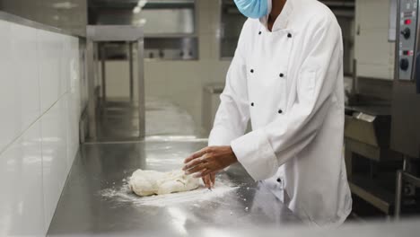 African-american-female-chef-wearing-face-masks-preparing-dough-in-restaurant-kitchen