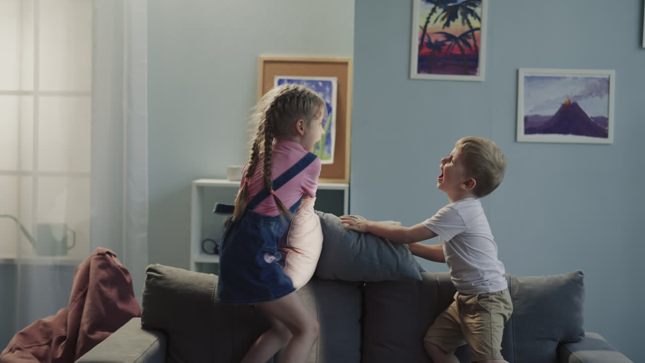 Laughing sibling children have pillow fight jumping on sofa