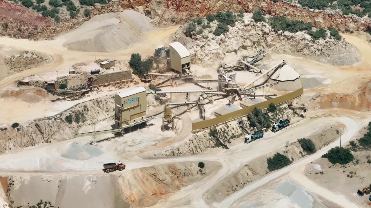 Aerial view of a cement production facility showing limestone quarry operations. Heavy machinery, conveyor belts, and dump trucks transport materials throughout the site.