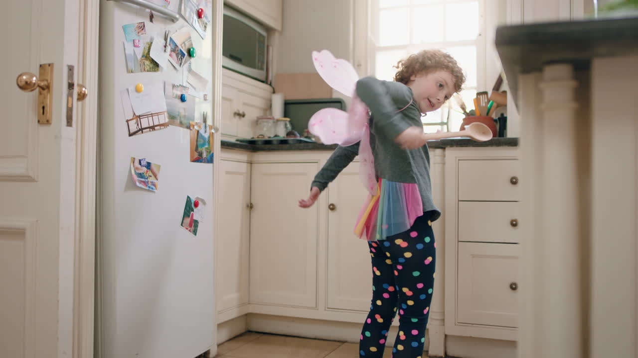 happy little girl dancing in kitchen wearing cute fairy wings having fun waving spoon playing pretend doing funny dance moves enjoying weekend at home