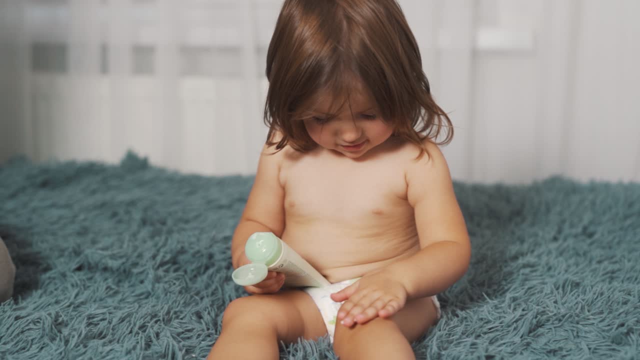 Una adorable niña de dos años con pañal se aplica crema en las piernas.