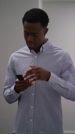 vertical video of young businessman in office looking at message with bad news on mobile phone and leaning against wall looking unhappy and depressed