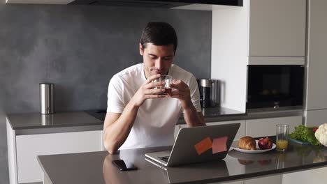 Portrait-of-a-handsome-relaxed-caucasian-man-in-a-white-shirt-sitting-on-kitchen-in-front-him-laptop-and-smartphone.-Guy-sipping-a-black-tea-from-transparent-mug-and-peacefully-smiling