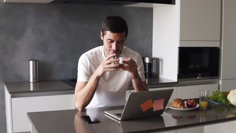 retrato de un apuesto hombre caucásico relajado con una camisa blanca sentado en la cocina delante de él portátil y teléfono inteligente. tipo bebiendo un té negro de una taza transparente y sonriendo pacíficamente