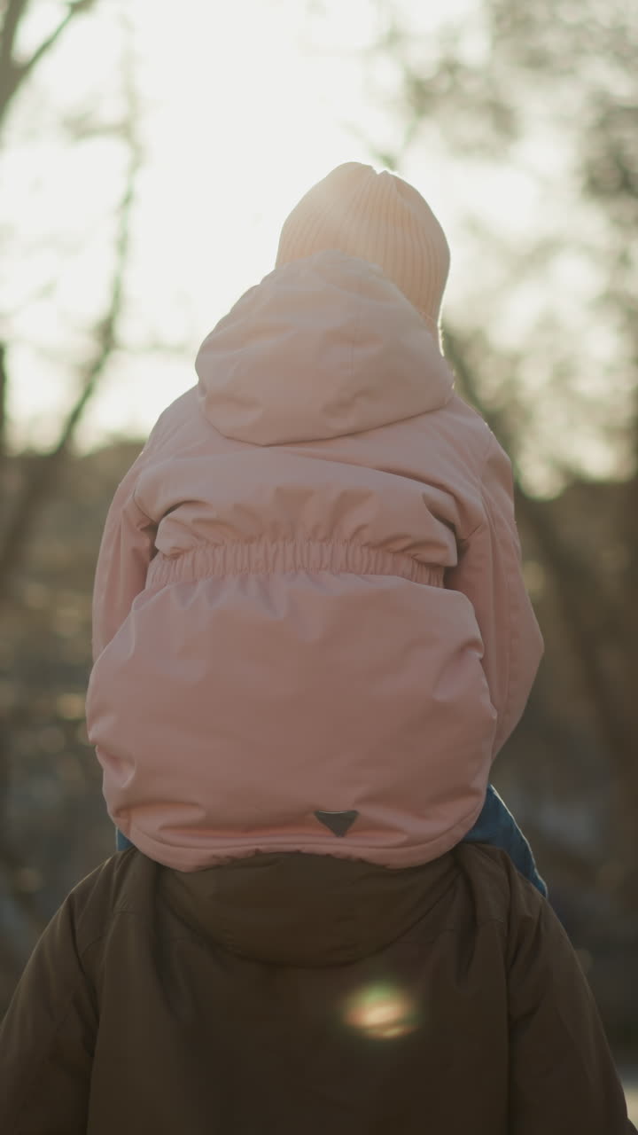 핑크색 모자와 재을 입은 어린 소녀가 공원을 걸어가는 동안 아버지의 목에 앉아있는 뒷면. 태양은 나무를 통해 빛나고 따뜻하고 조용한 분위기를 만니다.