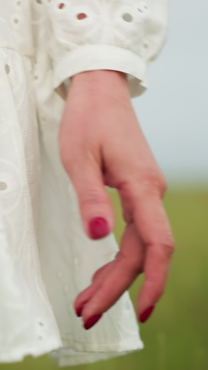 un primer plano de la mano de una mujer con esmalte de uñas rojo, moviéndose elegantemente a través de un campo de hierba borroso. ella lleva un vestido blanco con patrones, creando una atmósfera suave y serena en un entorno natural