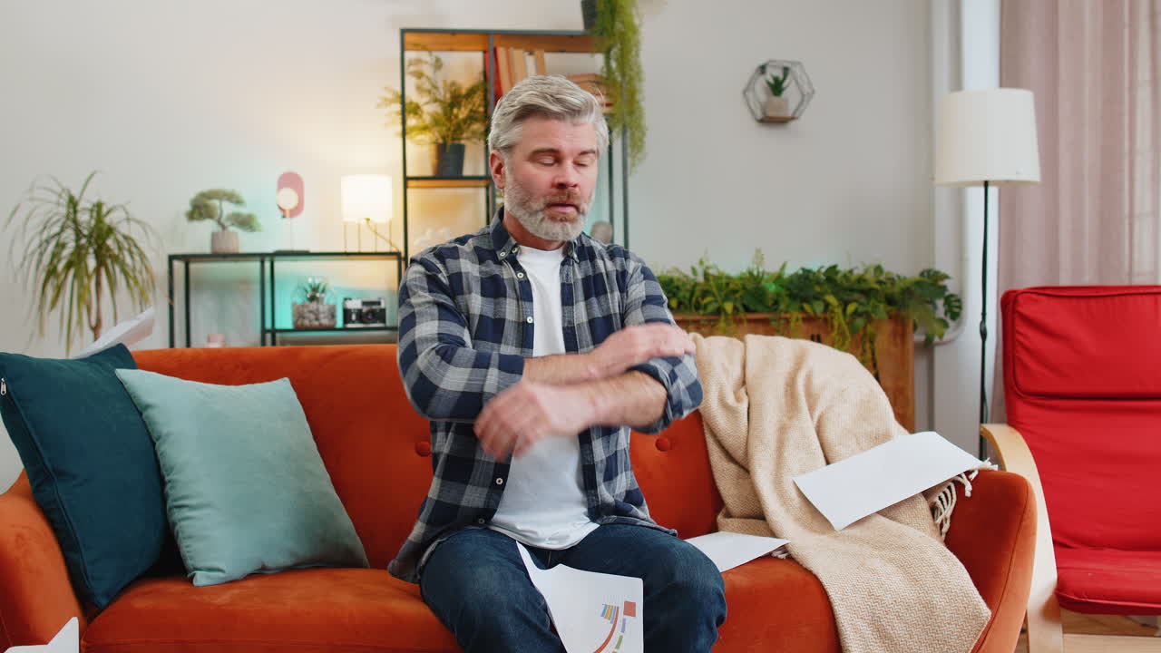 Dissatisfied senior man throwing out bills checks or papers documents while sitting on couch at home