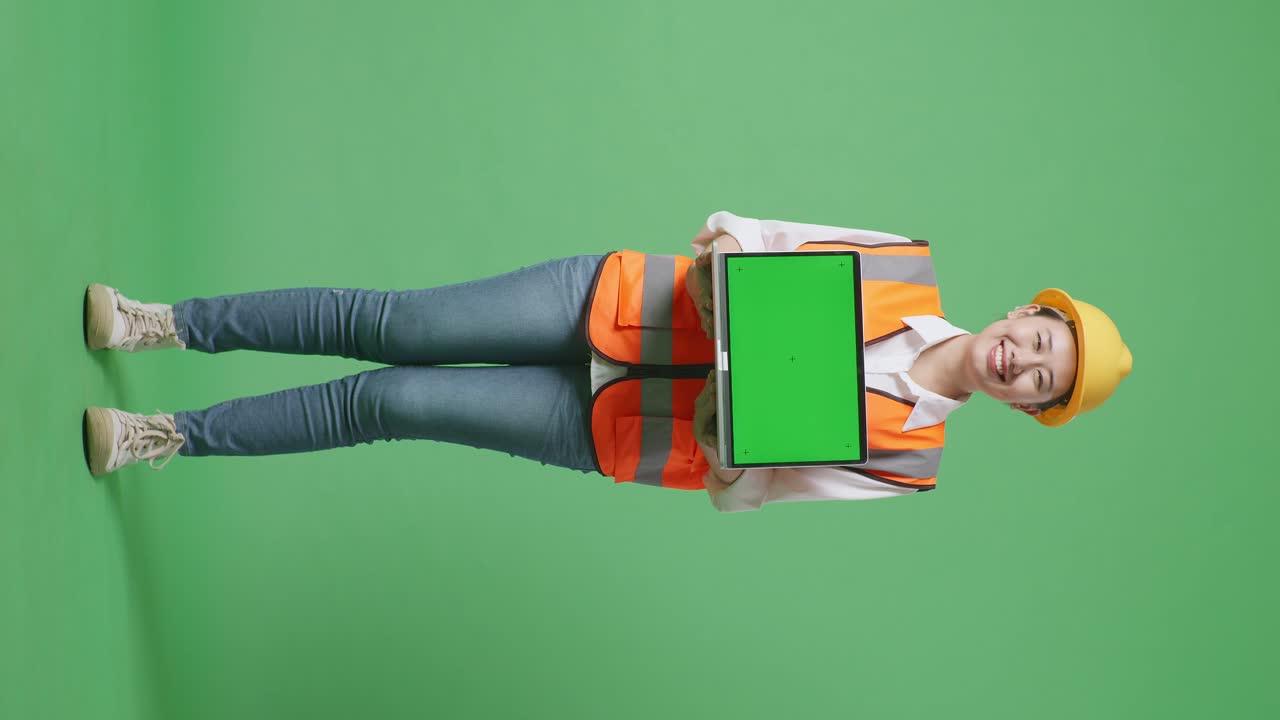 cuerpo lleno de ingeniera asiática con casco de seguridad sonriendo y mostrando portátil de pantalla verde a la cámara mientras está de pie en el estudio de fondo de pantalla verde