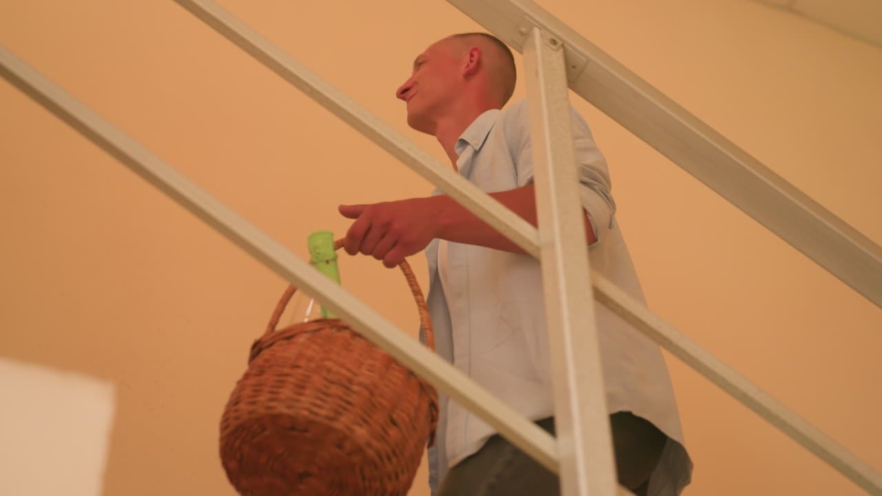 Kaukasisches Paar steigt mit Korb und Teddy die Treppe hoch und zieht in ein warmes Pfirsich-Loft ein. Der Mann führt mit einem Picknickkorb das Geländer hinauf, während die Frau mit einer kleinen Schachtel folgt, gemütliche häusliche Stimmung, lässig