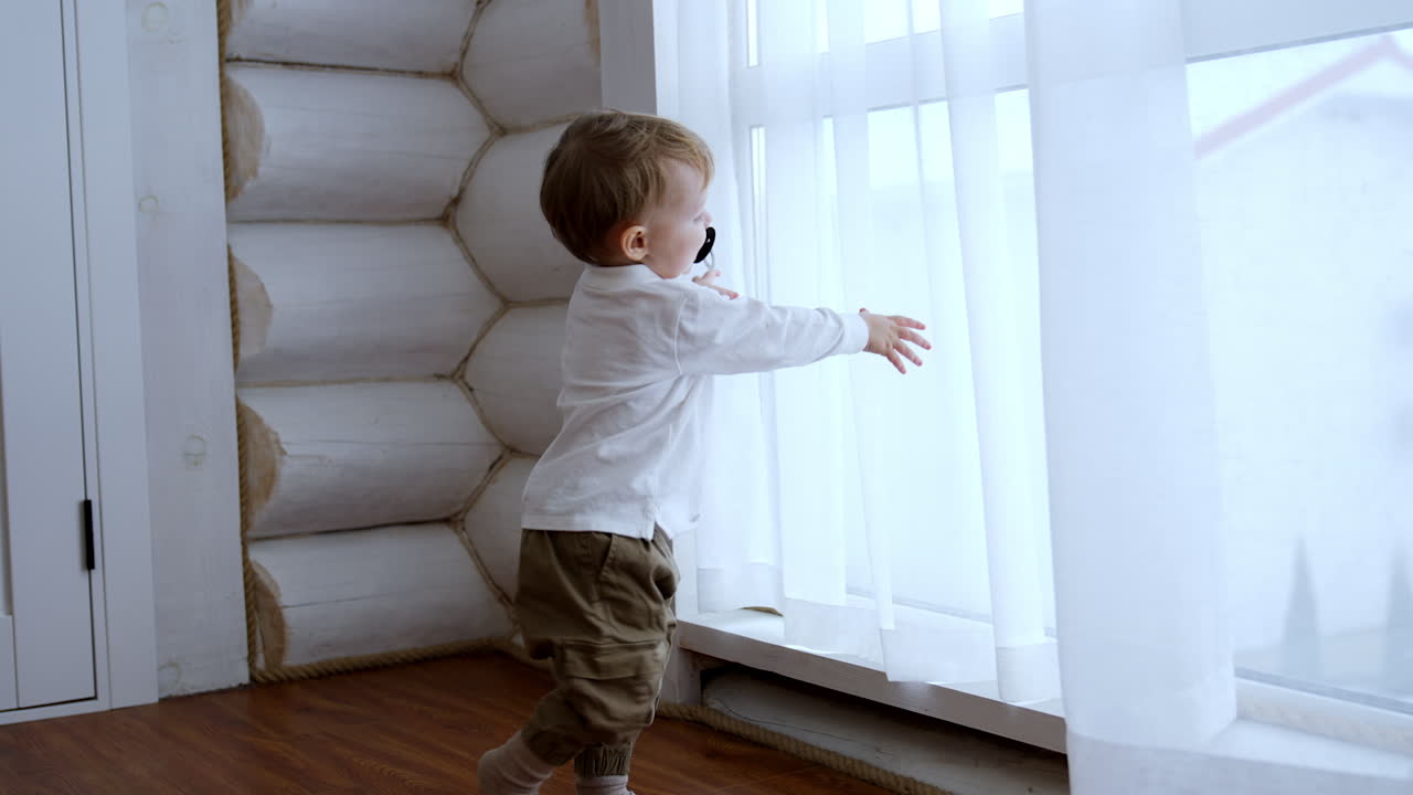 Adorable Caucasian toddler boy with a pacifier in mouth plays with white curtains. Happy kid playing cheerfully at the window.