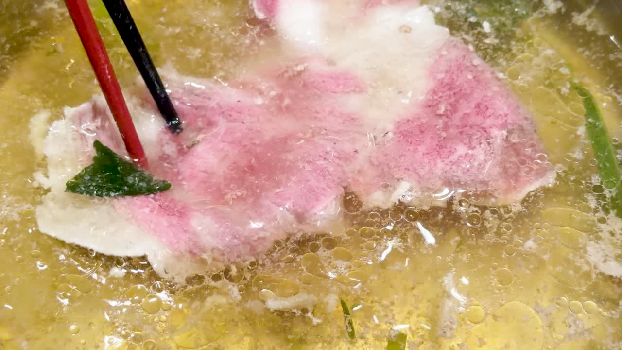 Marbled wagyu beef cooks in bubbling hotpot broth, held by chopsticks, under bright lighting